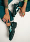 A person sitting on the floor, touching their LC0001 by Rafa teal and black low top sneakers matching their teal pants, with visible rings on their fingers.