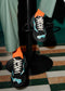 Partial view of a person's feet wearing black low top sneakers with white laces, orange and black socks, standing on a patterned green and white floor in LC0003 FreeYourMind.