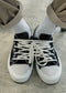 A close-up of V9 Antique-White & Lilac low top sneakers with untied laces, worn with white socks and tan pants.