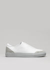 A V7 white leather slip-on sneaker with a plaster suede heel accent and a textured rubber sole, displayed against a neutral gray background.