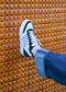 Wearing dark jeans and white sneakers handcrafted in Portugal, a person rests their foot on a textured orange wall, highlighting the black and purple accents of the ethically made N0019 by Ana Júlia.