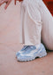 A person wearing L0024 by Bárbara sneakers, handcrafted from premium Italian leather in light blue and white, crouches on a pinkish surface, perfectly complemented by their white pants.