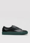The V49 Black w/ Forest Green is a Minimal Low slip-on sneaker featuring premium Italian leather, a dark green sole, and a subtle embossed logo on the side, handcrafted in Portugal against a plain light gray background.