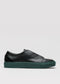 The V49 Black w/ Forest Green is a Minimal Low slip-on sneaker featuring premium Italian leather, a dark green sole, and a subtle embossed logo on the side, handcrafted in Portugal against a plain light gray background.