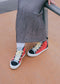 A person sits on a rail, wearing a wrinkled grey skirt and vibrant red and black TH0023 by Erica high-top sneakers with yellow laces, ethically handcrafted in Portugal.