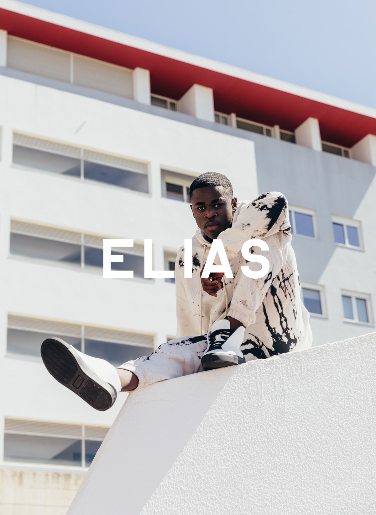 Elias seduto su un muro, indossando Diverge sneakers, promuovendo l'impatto sociale e le scarpe personalizzate attraverso il progetto Imagine.