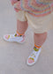 A person wearing TH0024 by Iara, vibrant high-top sneakers and socks, paired with beige shorts and a multicolored sweater, stands on a light gray surface. These premium canvas shoes are expertly handcrafted in Portugal for an artisanal touch.