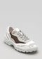 The L0021 by Hanin is a handcrafted sneaker in white, gray, and brown, featuring a chunky sole and lace-up design against a plain gray backdrop. It highlights impeccable craftsmanship and an ethically made commitment.