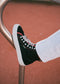 The TH0022 by João is a black multi-layered high-top sneaker with red accents and white laces, shown on a curved metal structure against a reddish background. Handcrafted in Portugal, it exemplifies fine craftsmanship and style.