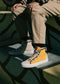 A person sits on steps wearing mismatched TH0020 by Leonardo sneakers, one beige and one yellow, paired with beige pants. Handcrafted in Portugal from premium canvas, these sneakers add a unique twist to the ensemble.
