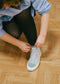 A person in a striped shirt ties the laces of a light blue TH0021 by Luana, an ethically made high-top sneaker, over black tights on a wooden floor.
