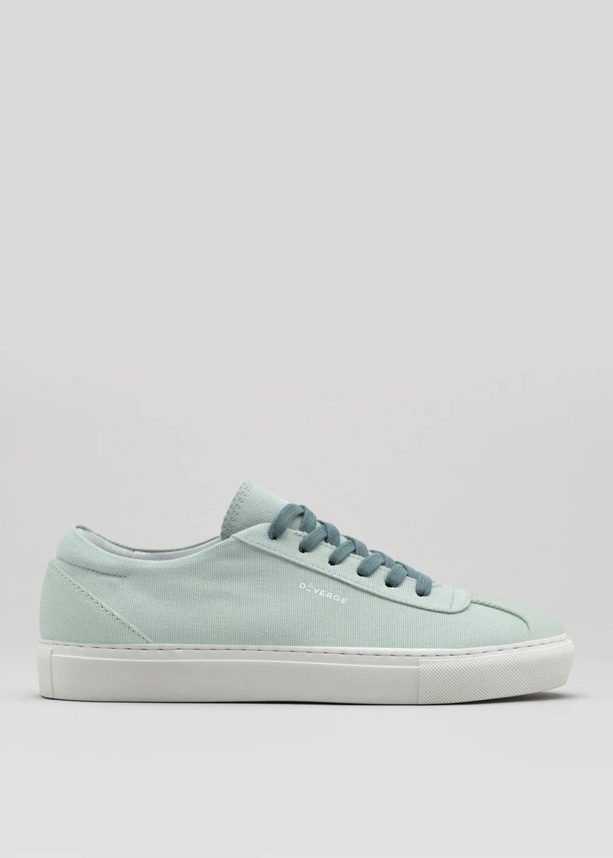 Introducing the V6 Sage Green, a versatile sneaker crafted with premium canvas featuring a light green hue, complemented by a white sole and blue laces against a plain background.