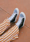 Person wearing TH0026 by Mafalda, black and white handcrafted sneakers with white laces, paired with orange and white striped pants on a textured brown surface.