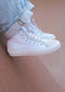 TH0025 by Nicole, handcrafted white high-top sneakers from Portugal, adorn a person's feet paired with light blue jeans slightly rolled up, standing on a sloped wooden surface.