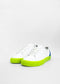 A pair of white slip-on sneakers with neon green soles and a blue accent on the heel, handcrafted in Portugal, against a white background.
