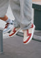 Two people sit on a rail in light-wash jeans and retro sneakers; one wears red and white, the other blue and white. These stylish M0024 by Rodrigo sneakers are crafted in Portugal, adding unique flair to their casual look.