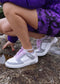 Una persona con un vestido morado se sienta en una roca junto al agua, mostrando su M0004 by Sara A. custom lavender and grey low top sneakers, con un pequeño tatuaje visible en su pierna izquierda.