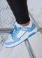 Gros plan du pied d'une personne sur une barre métallique, portant une paire de chaussures à la mode bleue et blanche M0002 by Sara Q sneakers avec une semelle épaisse.