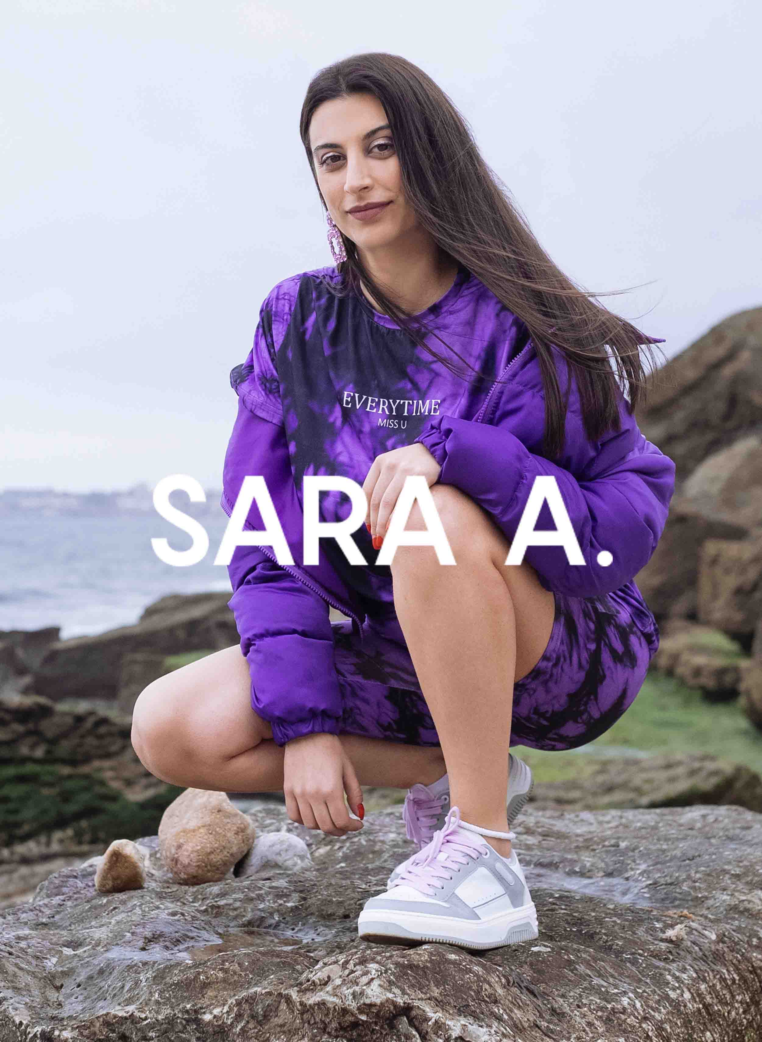 Sara indossa una camicia viola, abbinata a Diverge sneakers, mostrando le sue scarpe personalizzate e promuovendo l'impatto sociale attraverso il progetto Imagine.