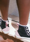 Close-up of a person wearing TH0016 by Ude, black and pink high-top sneakers with white laces. They are standing on a ledge with their legs visible. Handcrafted in Portugal, the premium canvas and soft suede shoes feature the brand logo on the rear.