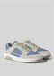 The V17 Lake Blue W/ Bone sneakers, handcrafted in Portugal with premium Italian leathers, feature a sleek retro-future design in grey and blue with white soles and laces on a minimalist grey background.