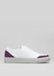 The V25 White W/ Plum is a handcrafted low-top sneaker made in Portugal from premium Italian leathers. It features a white design with plum accents on the toe and heel, a flat sole, and is set against a plain gray background.
