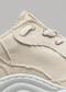 Primer plano de una zapatilla baja de color beige que muestra el tejido texturizado, los cordones y una parte de la suela blanca, con la palabra "V5 Full Color Antique White" visible.