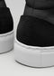 Nahaufnahme der Rückseite von schwarzen, strukturierten High-Top-Schuhen sneakers mit geprägtem Markenlogo und weißen Sohlen vor grauem Hintergrund. MH0003 von Chrys