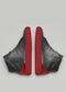 Un par de zapatos MH0010 Bred con parte superior de cuero negro y suela de color rojo brillante, vistos desde atrás sobre un fondo neutro.