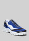 Un'unica sneaker low top multicolore con pannelli blu, neri e bianchi e suola bianca, su sfondo grigio. (L0004 di Daniel)