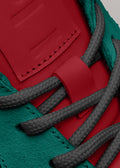 Nahaufnahme eines Low-Top-Sneakers L0010 Folie à Deux mit roten Leder- und türkisfarbenen Wildlederdetails und grauen, strukturierten Schnürsenkeln.