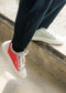 Phrase avec nom de produit : Deux hommes portant des chaussures basses de couleurs différentes sneakers; l'un TL0003 by Thierry en rouge et gris, l'autre en vert pâle, debout sur une surface en béton.