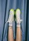V3 Ocean Blue Leather high-top sneakers with green soles worn with purple socks, feet crossed and placed against a blue curtain background.