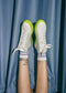 A pair of legs wearing V7 Grey Floater shoes with neon green soles and purple socks against a draped blue curtain background.