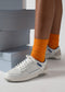 A person wearing M0004 by Sara A. low-top sneakers with blue accents and bright orange socks stands near stacks of gray-blue shoe boxes.