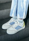 Eine Nahaufnahme der Füße einer Person, die elegante, niedrige V24 White & Electric Blue sneakers auf einem gestreiften Metallboden trägt.