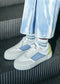 Primo piano dei piedi di una persona che indossa Now White Canvas low-top sneakers con pannelli personalizzati blu e bianchi, abbinati a jeans blu chiaro, in piedi su una superficie metallica a coste.