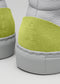 Detalle de la parte del talón de dos zapatos MH0005 Be Your Own Star de piel blanca con detalles de ante verde vibrante, con la marca en relieve.