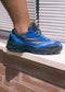 Close-up of a person's foot wearing a blue and black handcrafted L0015 by Iara with a chunky sole, resting on a white ledge against a background of brown bricks and white blinds.