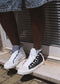 Person wearing mismatched black and white high-top sneakers, TH0017 by Khadija, handcrafted in Portugal, standing on a tiled surface near a glass door with blinds. Denim fabric is visible from the lower half of their clothing.