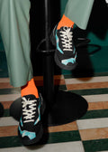 Ein Paar Füße mit leuchtend orangefarbenen Socken und schwarzen Maßschuhen mit weißen Schnürsenkeln, die auf einer V2 Color Mix Sage Green Basis vor einem grün-weiß gestreiften Boden stehen.