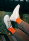 Un paio di DiVERGE X BUREL  Pearl low top sneakers abbinate a calzini arancioni brillanti e pantaloni grigi, appoggiati su un separé di pelle verde.