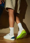 Person standing in a sunlit room wearing ML0036 White Leather W/ Yellow sneakers crafted with premium Italian leathers, featuring a green heel detail, white socks, and beige shorts.