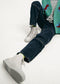 Person wearing denim jeans and grey high-top sneakers, with a MH0080 Green W/ Yellow cardigan featuring blue and red designs, sitting on a white background. Visible from the waist down.