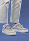 Una persona in piedi indossa jeans V9 White & Petrol Blue e una low top grigia sneakers su uno sfondo blu.