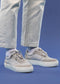 Close-up view of a person wearing grey N0004 Talent shoes and light blue jeans against a blue background.