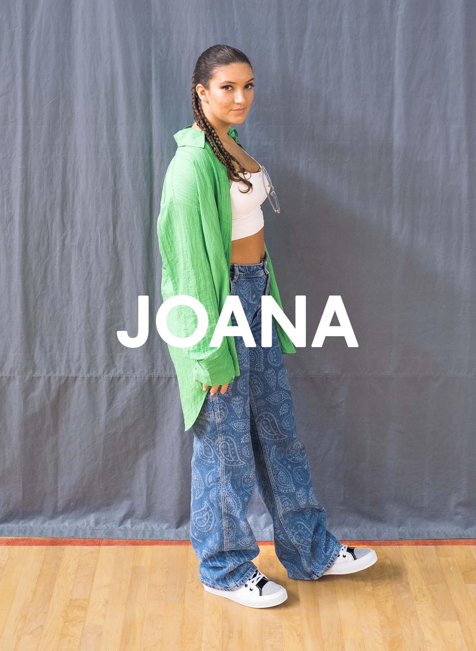 Joana con camisa verde y jeans de pie sobre un piso de madera, Diverge sneakers, promoviendo el impacto social y el calzado personalizado a través del proyecto Imagine.  