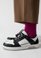 Nahaufnahme der Unterschenkel einer Person, die leuchtend rosafarbene Socken und M0002 by Sara Q low top sneakers trägt, gepaart mit einer braunen Hose vor einem weißen Hintergrund.