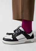 Persona de pie en V6 Arctic Blue W / White low top sneakers emparejado con calcetines de color rosa brillante y pantalones marrones sobre un fondo blanco.