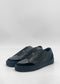 The SO0060 Black Floater, a pair of black slip-on sneakers crafted from premium Italian leathers with a smooth finish and black rubber soles, are displayed on a plain gray background.